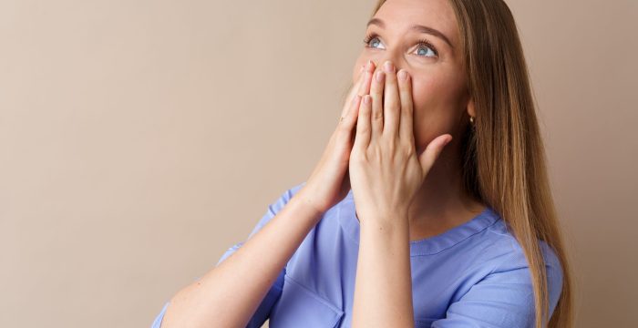 Shocked young woman looking up and .covers her mouth with hands against beige background